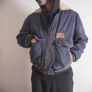 May include: A blue denim bomber jacket with a brown faux fur collar and a yellow, green, and black striped trim. The jacket has a zipper closure and a patch on the right chest that reads "Genuine Leather Garment Co. Made in USA".