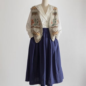 May include: A mannequin is dressed in a vintage ensemble. It features a white blouse with a lace collar, a grey knitted vest with floral and geometric designs, and a navy blue pleated skirt. The outfit is set against a white backdrop.