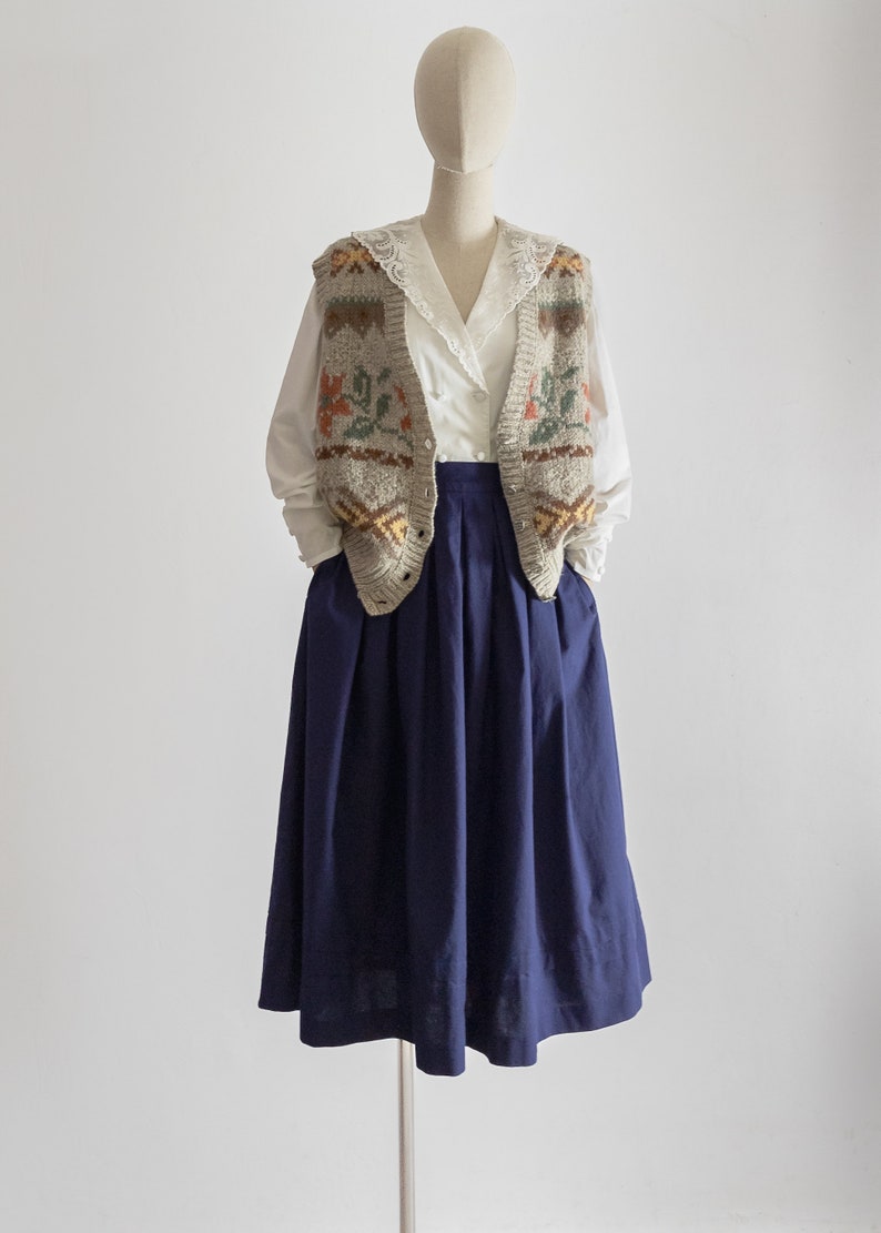 May include: A mannequin is dressed in a vintage ensemble. It features a white blouse with a lace collar, a grey knitted vest with floral and geometric designs, and a navy blue pleated skirt. The outfit is set against a white backdrop.