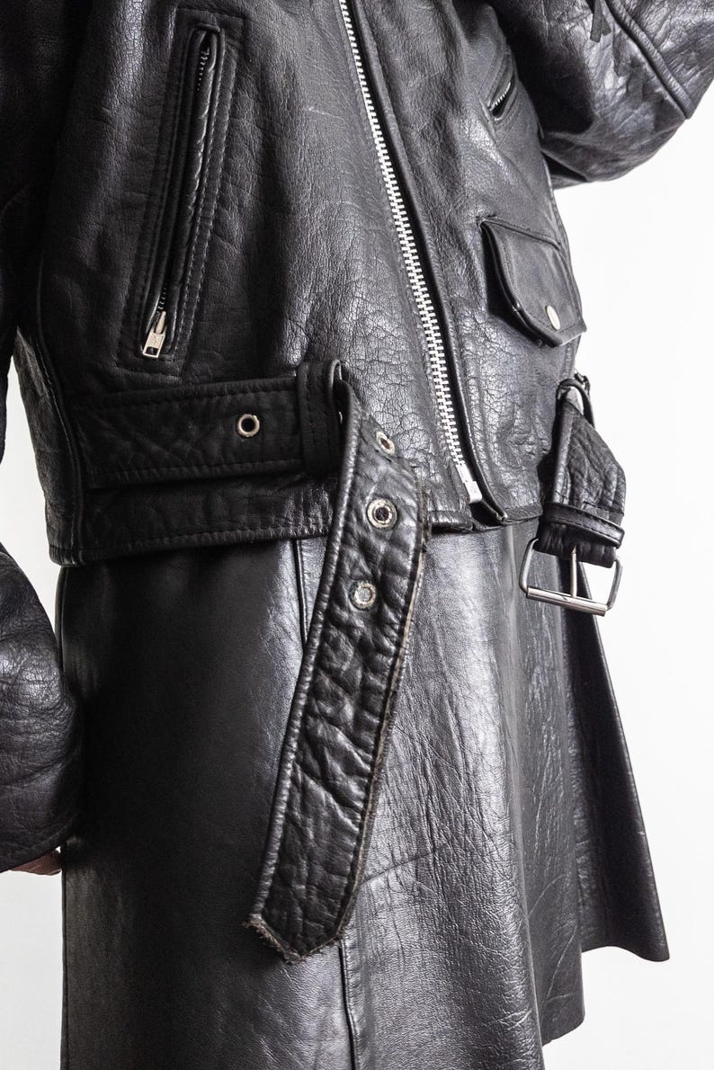 May include: Black leather jacket with a silver zipper, pockets, and belt details. The jacket has a classic biker style with a cropped length and a textured finish. The jacket features a silver buckle and metal rivets.