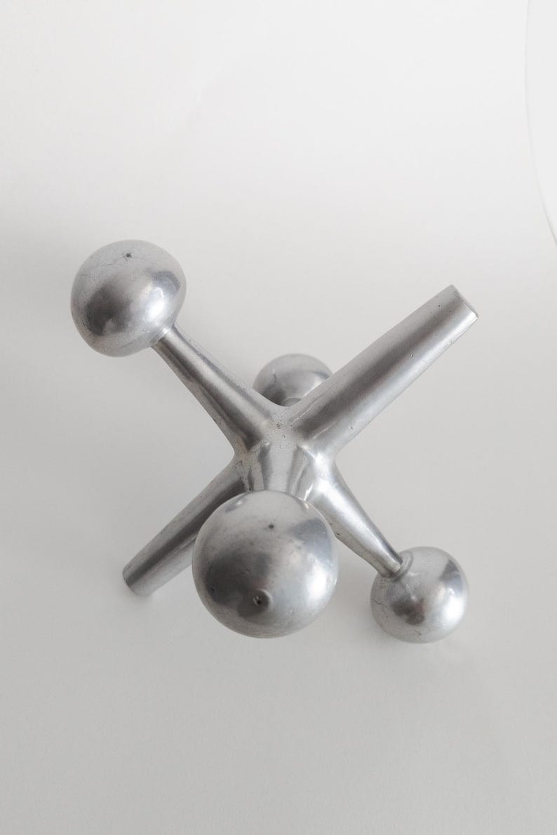 K&ouml;nnte beinhalten: Eine silberfarbene Metallskulptur in Form eines Springerspiels mit sechs strahlenf&ouml;rmigen Armen, die in glatten, kugelf&ouml;rmigen Enden auslaufen. Das Objekt hat eine matte, geb&uuml;rstete Metalloberfl&auml;che und eine zentrale Nabe, von der die Arme ausgehen.