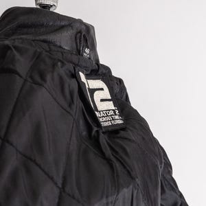 May include: Black quilted jacket with a leather collar. The jacket features a patch with the text "TERMINATOR 2 ACROSS TIME STUDIOS FLORIDA". The jacket is size 46.