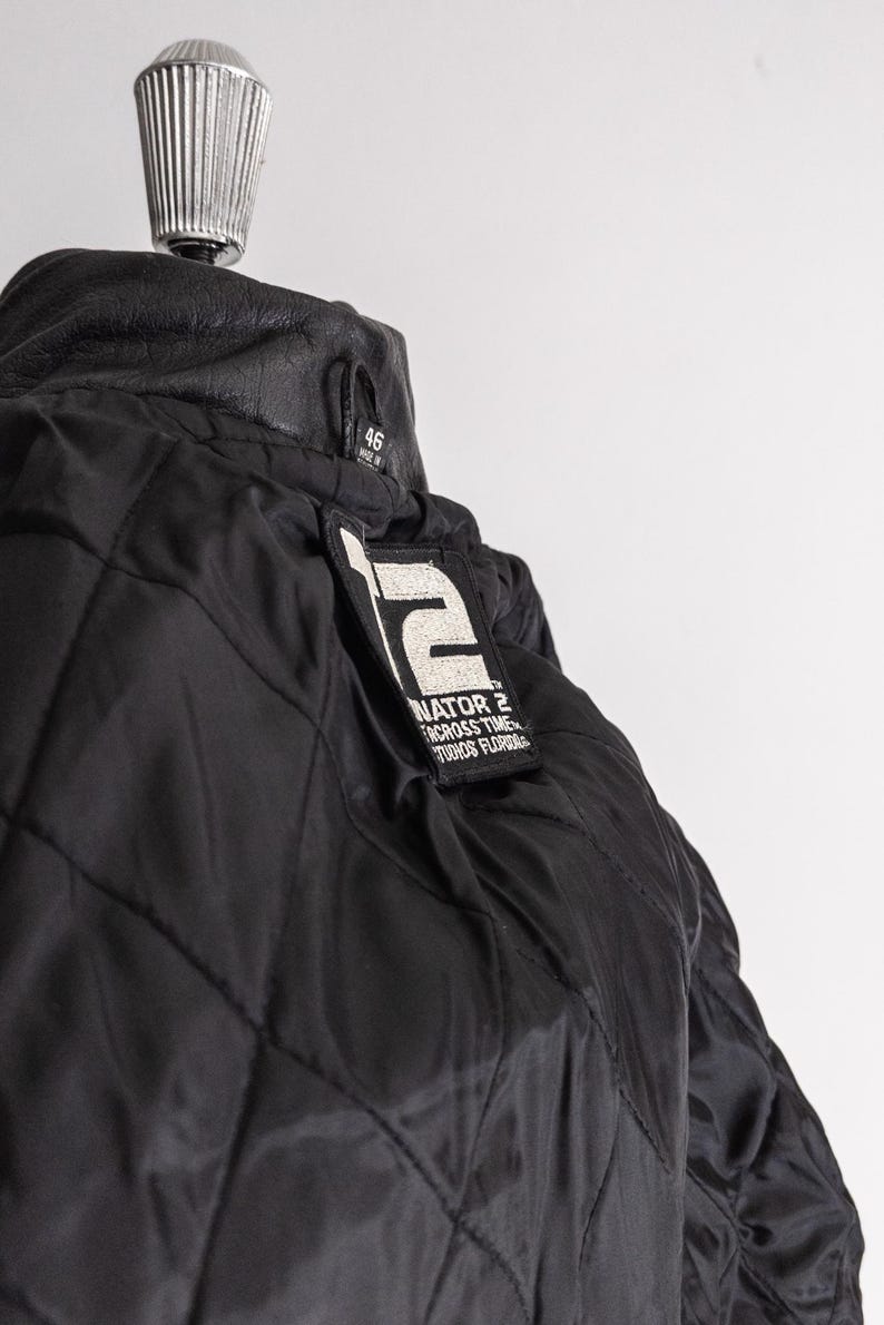 May include: Black quilted jacket with a leather collar. The jacket features a patch with the text "TERMINATOR 2 ACROSS TIME STUDIOS FLORIDA". The jacket is size 46.