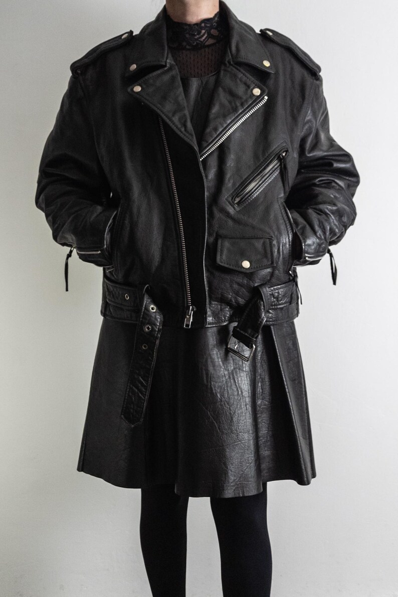 May include: Black leather biker jacket with a matching skirt. The jacket features a diagonal zipper, multiple pockets, and a belted waist. The skirt is a flared style with a buckle belt. The outfit is completed with black leggings.