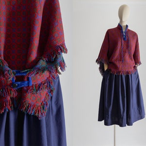 Op de afbeelding: Een rood en blauw gepatroneerd poncho met franjes, gedragen over een donkerblauwe rok. De poncho heeft een strik sluiting aan de hals.