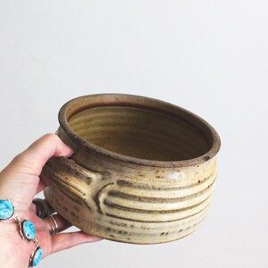 May include: A handmade ceramic bowl with a handle. The bowl is a light brown color with horizontal ridges around the exterior. The rim and handle are a darker brown. The bowl is held by a hand.