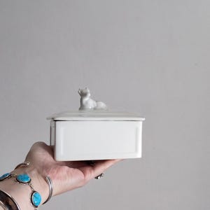 Può includere: Una scatola rettangolare bianca in ceramica con coperchio. Una piccola statuina bianca di un animale reclinato è sopra il coperchio. La scatola è tenuta in mano su uno sfondo grigio.