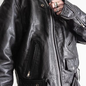 May include: Black leather jacket with a high collar, multiple pockets, and a front zipper. The jacket has a textured appearance and features silver hardware, including zippers and snaps. The design includes shoulder epaulets and a belt.