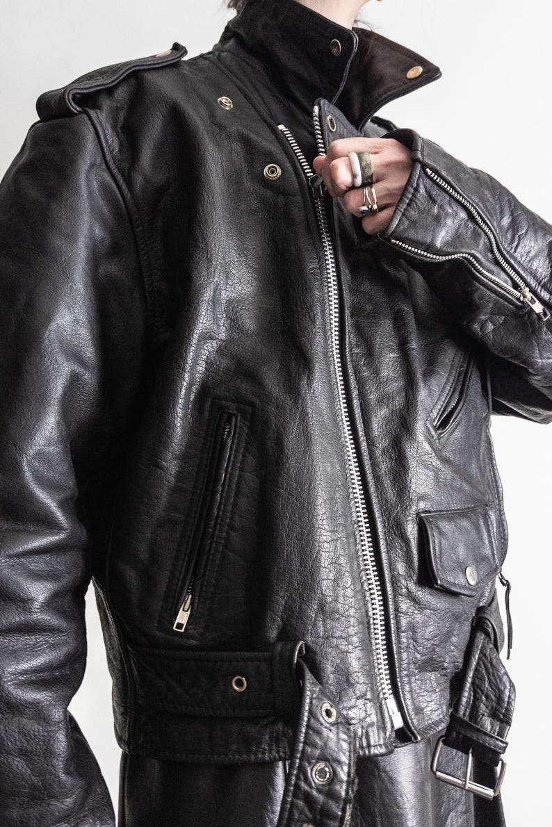 May include: Black leather jacket with a high collar, multiple pockets, and a front zipper. The jacket has a textured appearance and features silver hardware, including zippers and snaps. The design includes shoulder epaulets and a belt.