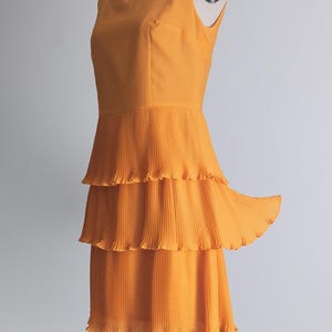 May include: A sleeveless, orange dress with a square neckline and a tiered, pleated skirt. The dress is displayed on a mannequin against a plain white background. The dress has a fitted bodice and a flowing skirt.