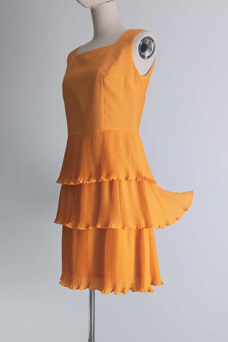 May include: A sleeveless, orange dress with a square neckline and a tiered, pleated skirt. The dress is displayed on a mannequin against a plain white background. The dress has a fitted bodice and a flowing skirt.