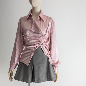 May include: A light pink, long-sleeved blouse with a wrap-style front and ruched detailing. The blouse has a collared neckline and is paired with a gray pleated skirt. The outfit is displayed on a mannequin.