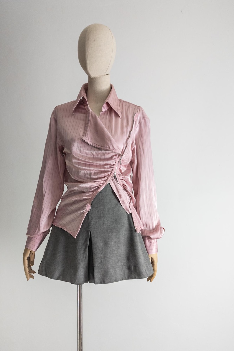 May include: A light pink, long-sleeved blouse with a wrap-style front and ruched detailing. The blouse has a collared neckline and is paired with a gray pleated skirt. The outfit is displayed on a mannequin.