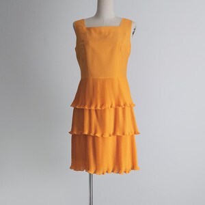 May include: An orange sleeveless dress with a square neckline and a tiered, pleated skirt. The dress is displayed on a mannequin against a plain white background. The dress is a vibrant shade of orange.
