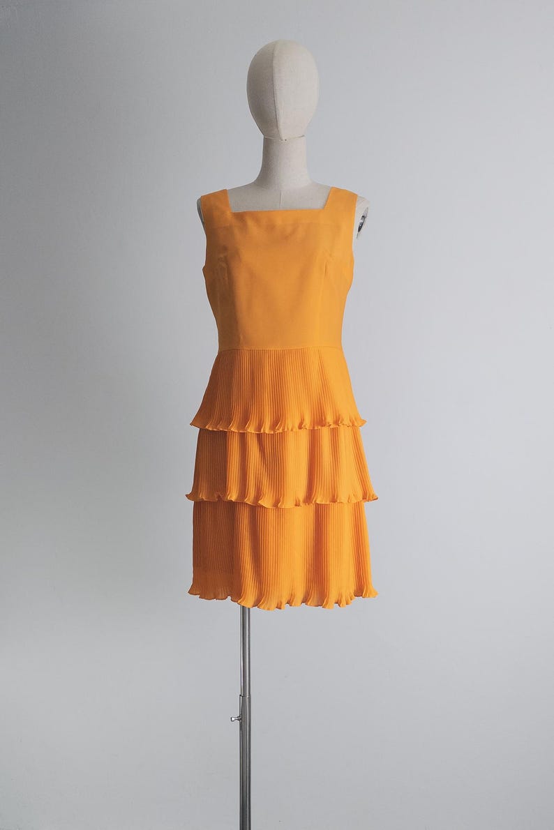 May include: An orange sleeveless dress with a square neckline and a tiered, pleated skirt. The dress is displayed on a mannequin against a plain white background. The dress is a vibrant shade of orange.