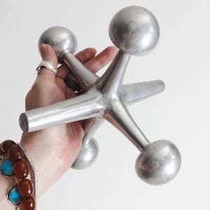 K&ouml;nnte beinhalten: Eine gro&szlig;e, silberfarbene Metallskulptur in Form eines Spielzeug-Jacks mit einem zentralen Knotenpunkt und sechs strahlenf&ouml;rmigen Armen, die jeweils in einer glatten, abgerundeten Kugel enden. Das Objekt hat eine matte Metalloberfl&auml;che.