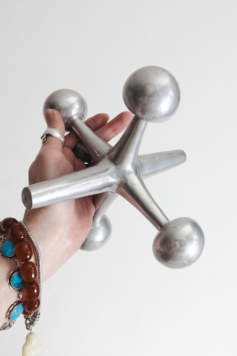 K&ouml;nnte beinhalten: Eine gro&szlig;e, silberfarbene Metallskulptur in Form eines Spielzeug-Jacks mit einem zentralen Knotenpunkt und sechs strahlenf&ouml;rmigen Armen, die jeweils in einer glatten, abgerundeten Kugel enden. Das Objekt hat eine matte Metalloberfl&auml;che.