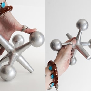 K&ouml;nnte beinhalten: Ein gro&szlig;es, silberfarbenes Metallspielzeug in Form eines Jacks mit sechs strahlenf&ouml;rmigen Armen, die jeweils in einer glatten, kugelf&ouml;rmigen Kugel enden. Das Objekt hat eine matte, metallische Oberfl&auml;che und eine geometrische, sternartige Struktur.
