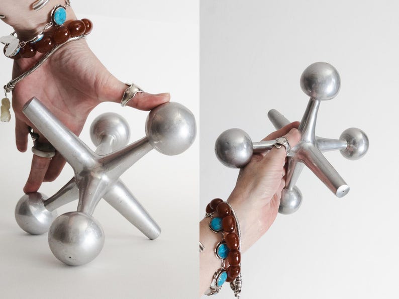K&ouml;nnte beinhalten: Ein gro&szlig;es, silberfarbenes Metallspielzeug in Form eines Jacks mit sechs strahlenf&ouml;rmigen Armen, die jeweils in einer glatten, kugelf&ouml;rmigen Kugel enden. Das Objekt hat eine matte, metallische Oberfl&auml;che und eine geometrische, sternartige Struktur.