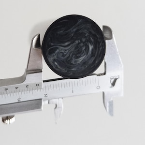 Könnte beinhalten: Ein runder, schwarz-weiß marmorierter Knopf, der in den Backen eines Metallmessschiebers gehalten wird. Das Messwerkzeug zeigt, dass der Knopf einen Durchmesser von etwa 3,8 cm hat. Die Oberfläche des Knopfes weist ein wirbelndes Muster aus Dunkelgrau und Weiß auf.