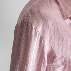 May include: Close-up of a pink button-down shirt with thin, vertical black stripes. The fabric appears to be a smooth, possibly satin-like material. The collar and shoulder detail are visible.