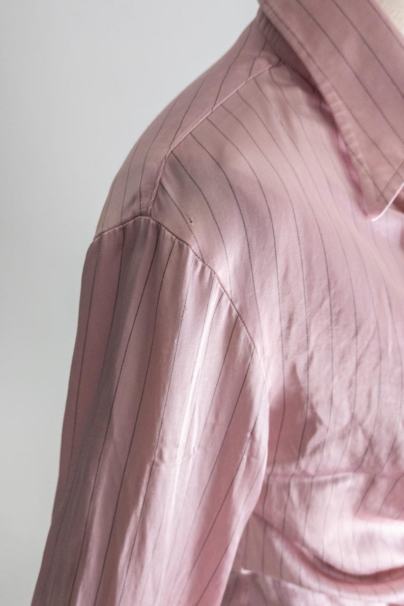 May include: Close-up of a pink button-down shirt with thin, vertical black stripes. The fabric appears to be a smooth, possibly satin-like material. The collar and shoulder detail are visible.