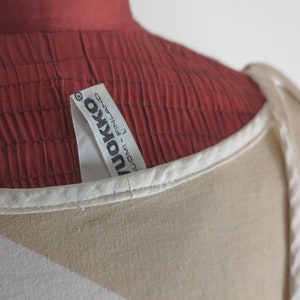 Op de afbeelding: Witte top zonder mouwen met een bruin en wit geometrisch patroon. De top heeft een wit label met de tekst "Ukonva - FINLAND" erop.
