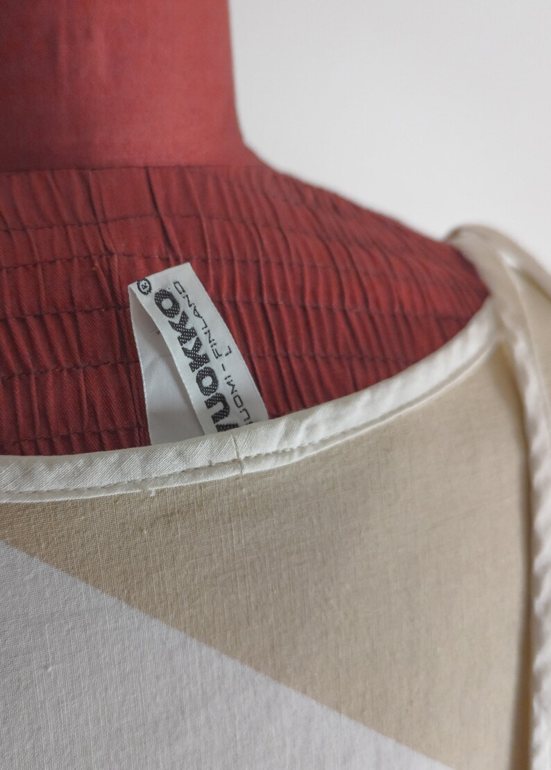Op de afbeelding: Witte top zonder mouwen met een bruin en wit geometrisch patroon. De top heeft een wit label met de tekst "Ukonva - FINLAND" erop.