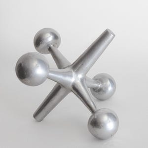 K&ouml;nnte beinhalten: Eine silberfarbene Metallskulptur in Form eines Spielzeug-Jacks mit sechs strahlenf&ouml;rmigen Armen, die jeweils in einer glatten Kugel enden. Das Objekt hat eine matte Metalloberfl&auml;che und eine geometrische, multidirektionale Struktur auf drei Kugeln.