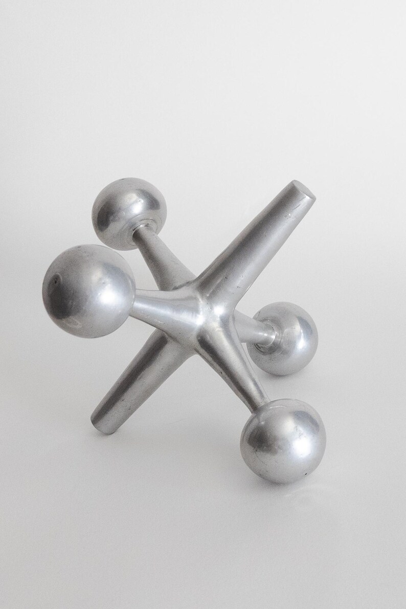 K&ouml;nnte beinhalten: Eine silberfarbene Metallskulptur in Form eines Spielzeug-Jacks mit sechs strahlenf&ouml;rmigen Armen, die jeweils in einer glatten Kugel enden. Das Objekt hat eine matte Metalloberfl&auml;che und eine geometrische, multidirektionale Struktur auf drei Kugeln.