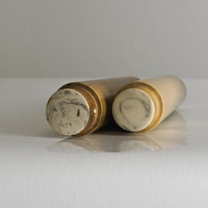 Könnte beinhalten: Zwei zylindrische Objekte in hellen Beige- und Goldtönen. Die Enden der Zylinder weisen ein marmoriertes weißes und graues Muster auf. Sie liegen auf einer weißen Oberfläche. Die Objekte scheinen dekorativ oder funktional zu sein.