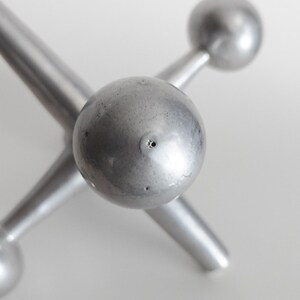 K&ouml;nnte beinhalten: Eine silberfarbene Metallskulptur in Form eines Spiel-Jacks mit sechs strahlenf&ouml;rmigen Armen, die in glatten, kugelf&ouml;rmigen Enden auslaufen. Die Metalloberfl&auml;che weist eine subtile, matte Textur mit kleinen, sichtbaren Unregelm&auml;&szlig;igkeiten auf.
