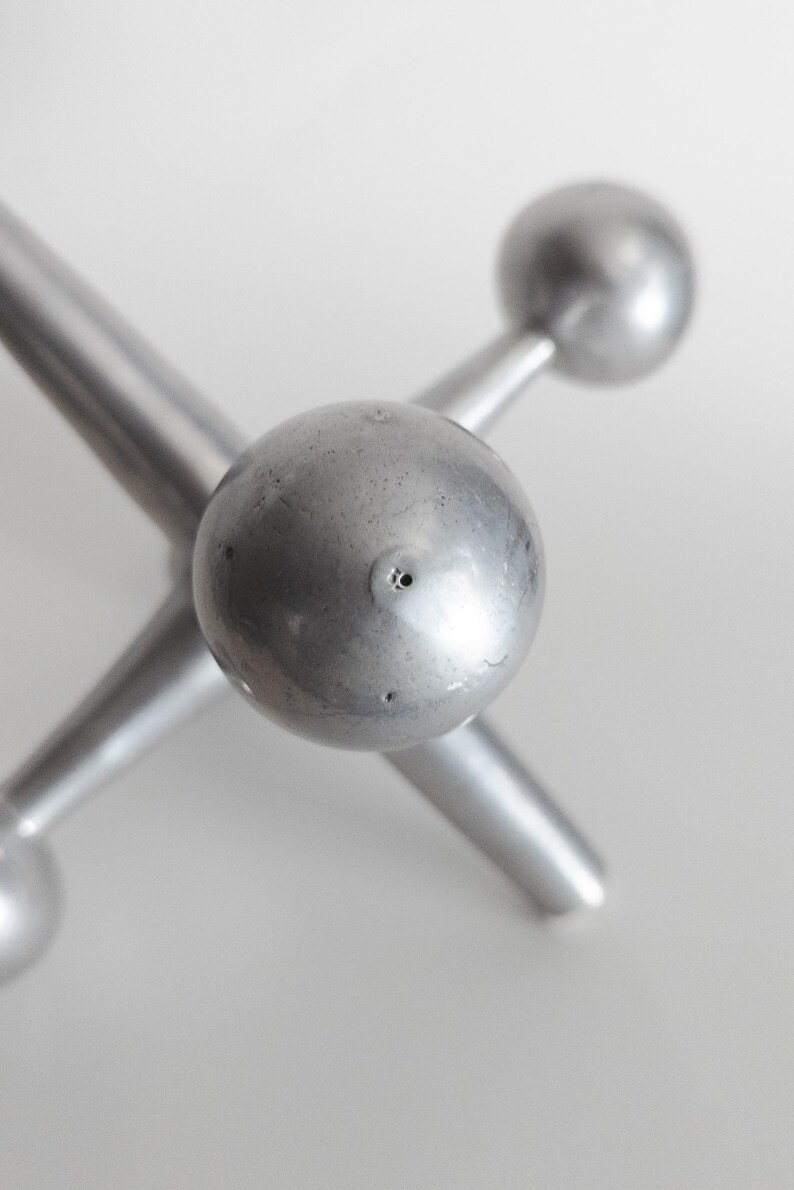 K&ouml;nnte beinhalten: Eine silberfarbene Metallskulptur in Form eines Spiel-Jacks mit sechs strahlenf&ouml;rmigen Armen, die in glatten, kugelf&ouml;rmigen Enden auslaufen. Die Metalloberfl&auml;che weist eine subtile, matte Textur mit kleinen, sichtbaren Unregelm&auml;&szlig;igkeiten auf.