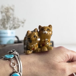 Peut inclure: Deux petites figurines décoratives de chats. Les chats sont bruns avec des rayures noires et des accents colorés. Ils sont assis côte à côte. Un bracelet en argent avec des pierres turquoise est visible au premier plan.