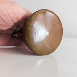 Puede incluir: Primer plano de un objeto circular de color bronce, con una parte superior ligeramente abovedada. La superficie tiene un patrón texturizado y concéntrico que irradia desde el centro. El objeto está sostenido por una mano, lo que sugiere que es un objeto pequeño y portátil.