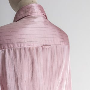 May include: A light pink button-down shirt with thin, vertical black stripes. The shirt is displayed on a mannequin, showcasing the back and collar details. The fabric has a subtle sheen, suggesting a satin or silk material.