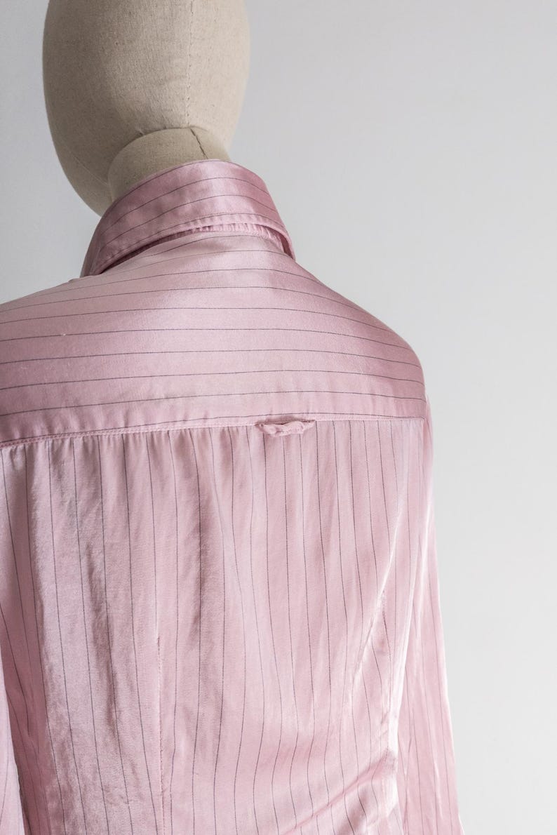 May include: A light pink button-down shirt with thin, vertical black stripes. The shirt is displayed on a mannequin, showcasing the back and collar details. The fabric has a subtle sheen, suggesting a satin or silk material.