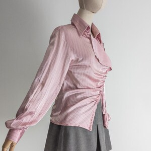 May include: A light pink, long-sleeved blouse with vertical pinstripes. The blouse features a collared neckline and a ruched detail on the side. It is paired with a gray pleated skirt.