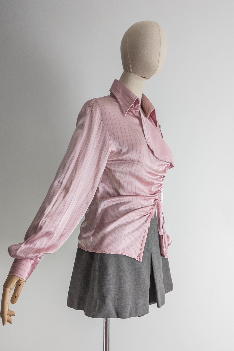 May include: A light pink, long-sleeved blouse with vertical pinstripes. The blouse features a collared neckline and a ruched detail on the side. It is paired with a gray pleated skirt.