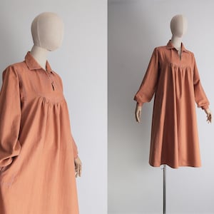 Puede incluir: Vestido color melocotón de manga larga, cuello camisero y detalle de ojo de cerradura. El vestido presenta un corpiño fruncido y un corte holgado, expuesto en un maniquí. Las mangas terminan en puños. El vestido es de tejido suave.