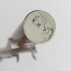 Puede incluir: Un objeto cilíndrico con un extremo liso de color crema que presenta un patrón de mármol negro en espiral. El cuerpo del objeto es de un tono marrón cálido y está colocado junto a una herramienta de medición metálica de 1,3 centímetros.