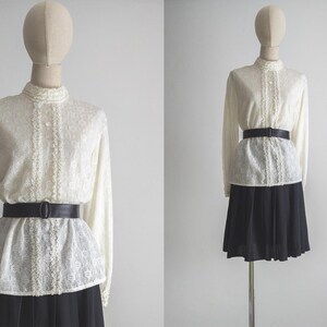 Op de afbeelding: Crèmekleurige blouse met lange mouwen, een hoge kraag en kanten details. De blouse wordt gecombineerd met een zwarte riem en een zwarte rok. De etalagepop wordt gepresenteerd tegen een effen witte achtergrond.