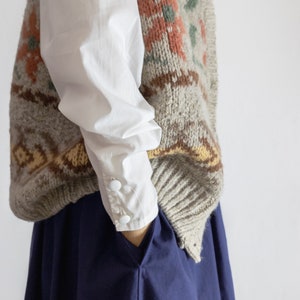 May include: A knitted wool vest with a floral pattern in shades of orange, green, and brown over a white long-sleeved shirt. The vest is layered over a navy blue skirt. The shirt has buttoned cuffs.