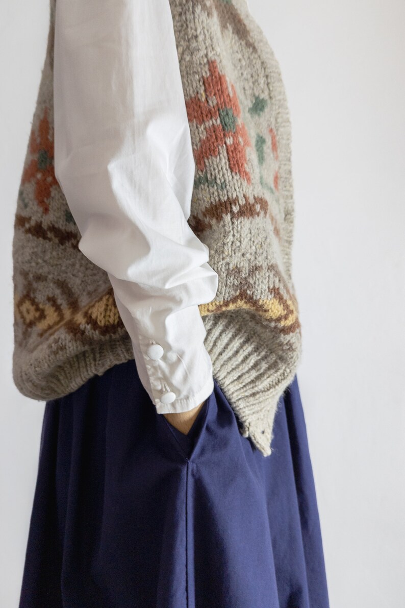 May include: A knitted wool vest with a floral pattern in shades of orange, green, and brown over a white long-sleeved shirt. The vest is layered over a navy blue skirt. The shirt has buttoned cuffs.