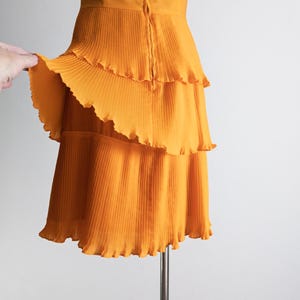 May include: A sleeveless, orange dress with a tiered, pleated skirt. The dress has a fitted bodice and a zipper closure down the back. The fabric has a slight sheen, and the hemline is scalloped.
