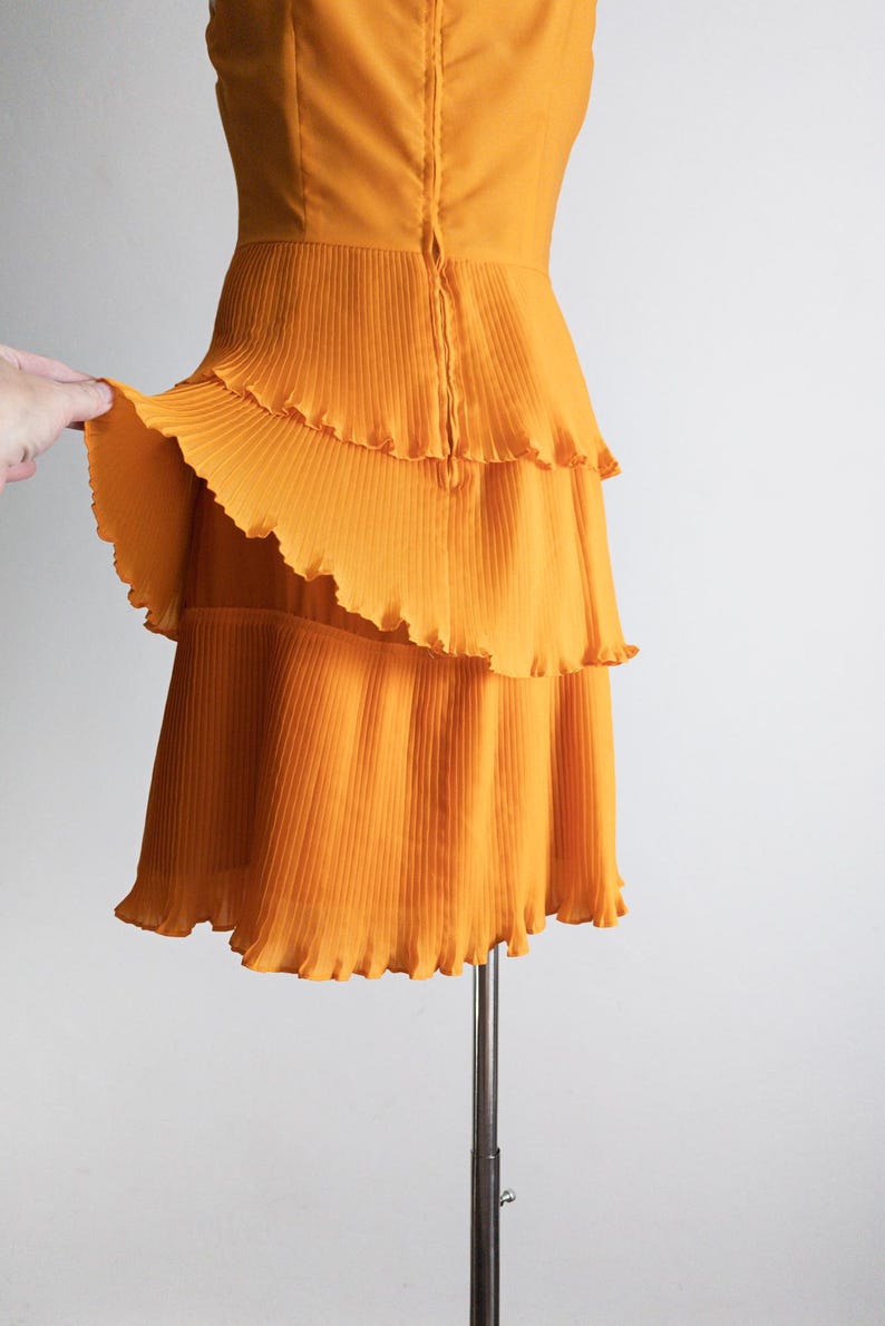 May include: A sleeveless, orange dress with a tiered, pleated skirt. The dress has a fitted bodice and a zipper closure down the back. The fabric has a slight sheen, and the hemline is scalloped.