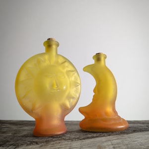 May include: Two glass bottles shaped like the sun and moon. The sun bottle is yellow and orange with a face and rays. The moon bottle is yellow and orange with a crescent moon shape.