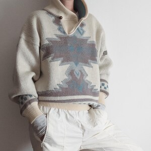 May include: A cream-coloured knit jumper featuring a large geometric pattern in shades of brown and blue across the chest. The jumper has a high collar with a single button closure, long sleeves with ribbed cuffs, and a ribbed hemline.