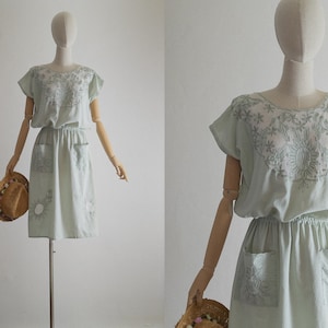 Könnte beinhalten: Ein hellgrünes Kleid mit kurzen Ärmeln und einem floralen Stickmuster. Das Kleid hat eine geraffte Taille und zwei Taschen auf der Vorderseite.