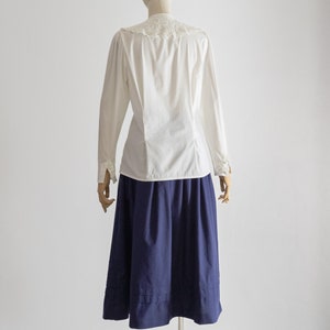 May include: A white blouse with a lace collar and long sleeves, paired with a navy blue skirt. The skirt is a knee-length A-line design. The outfit is displayed on a mannequin, showcasing a classic style.